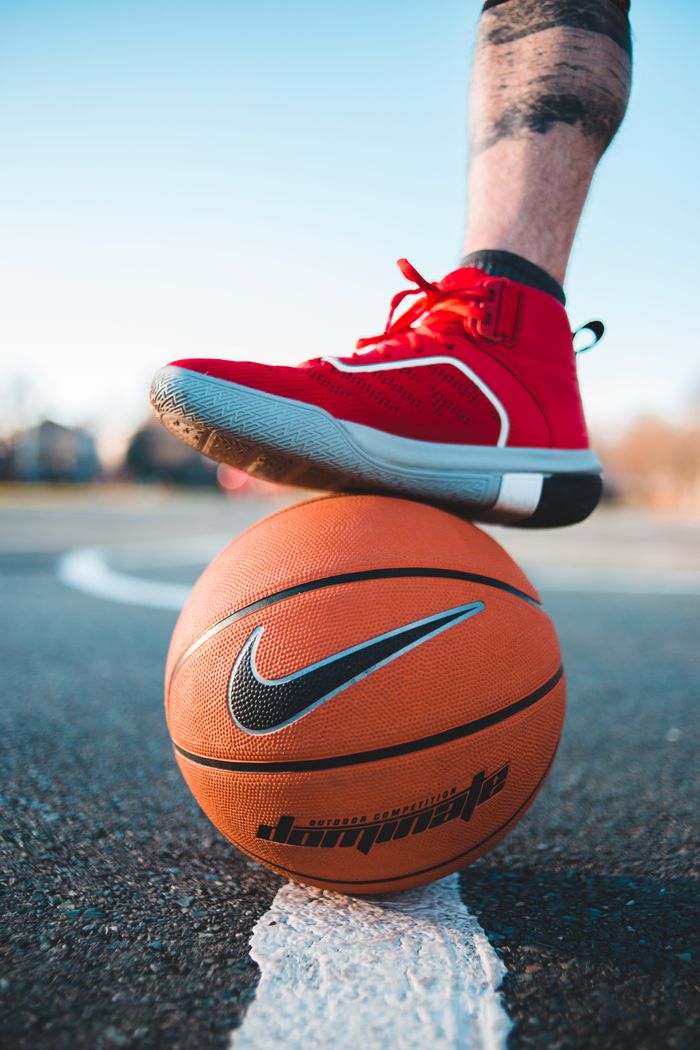 Crop faceless sportive man in red sneakers with foot on orange basketball placed on asphalt sports ground during training on street
